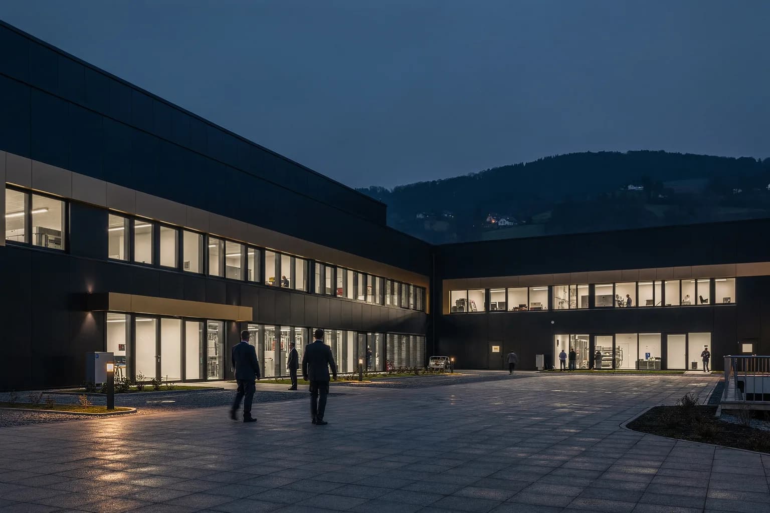 Moderner Technikbetrieb in der Dämmerung mit langen beleuchteten Fensterbändern und aufgeräumtem Vorplatz, Hügel der Bergstraße dahinter