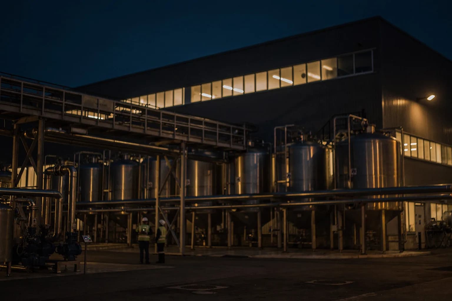 Großes Lebensmittelwerk in der Dämmerung mit Edelstahltanks im Freien, Rohrbrücken und beleuchteten Fensterbändern