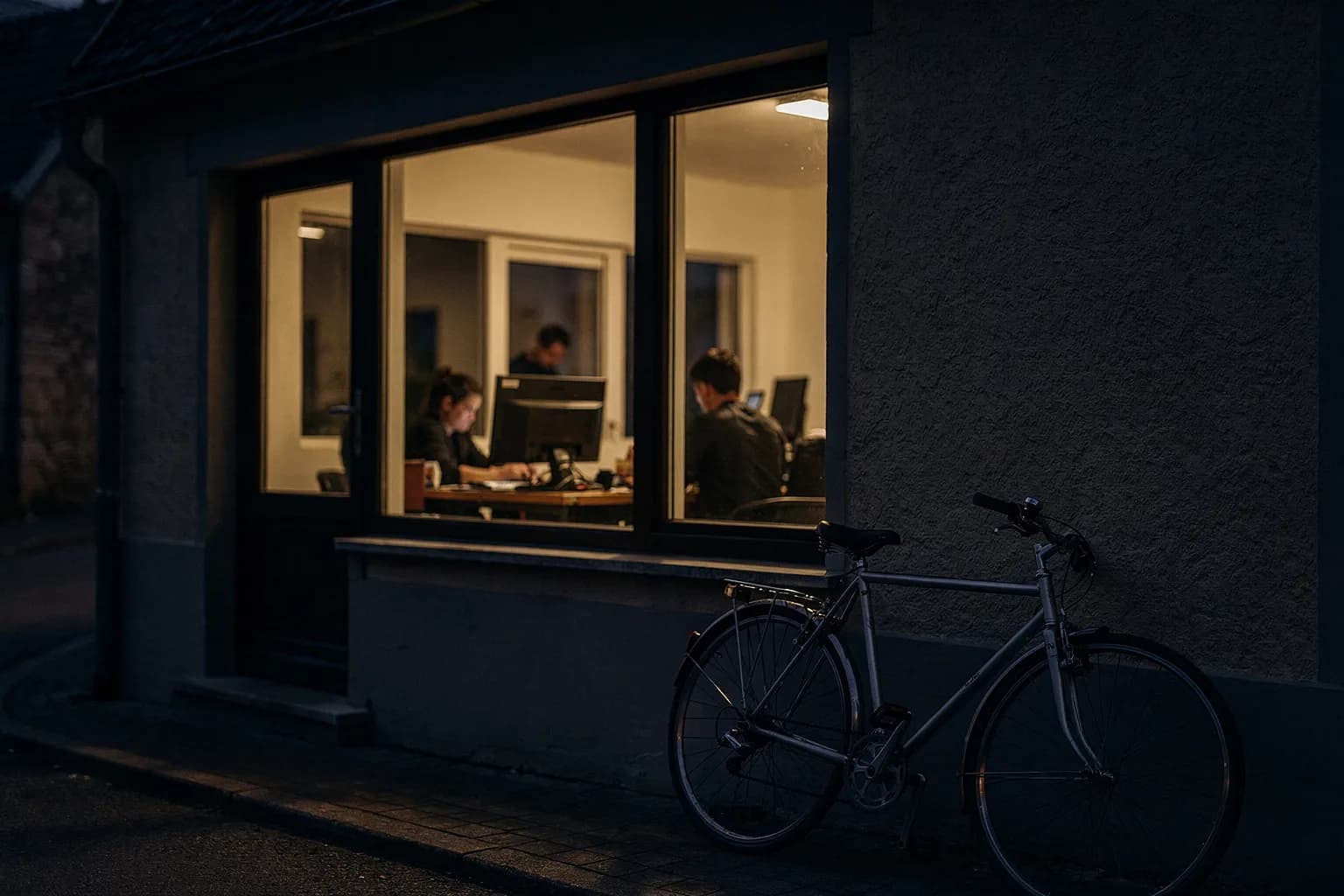 Kleines Büro in einem Dorf in der Dämmerung mit warm beleuchteten Fenstern und einem an der Wand lehnenden Fahrrad