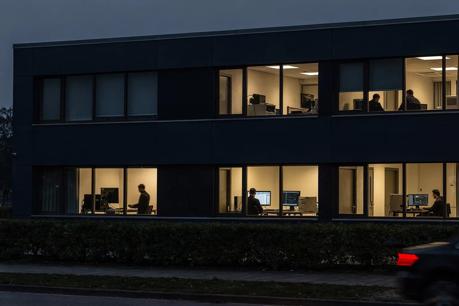 Schlichtes Technikgebäude in der Dämmerung mit beleuchteten Bürofenstern und einer geschnittenen Hecke am Gehweg