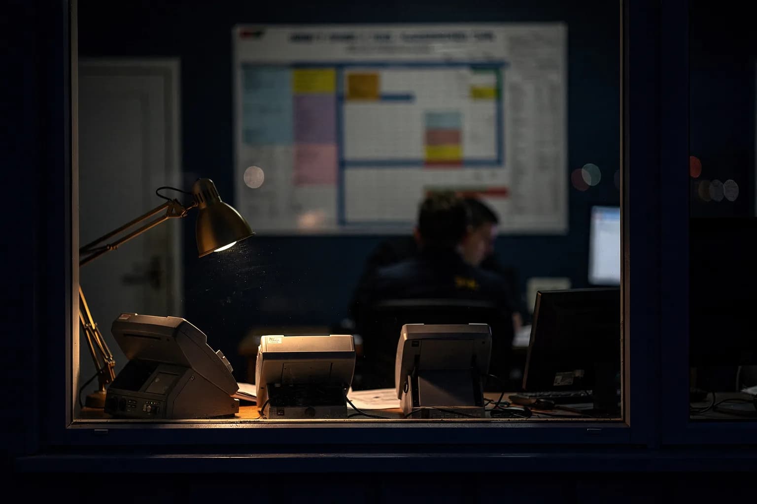 Fenster eines Hafenbüros bei Nacht, im Inneren eine Wandtafel mit Liegeplätzen, warme Lampe gegen den dunklen Kai
