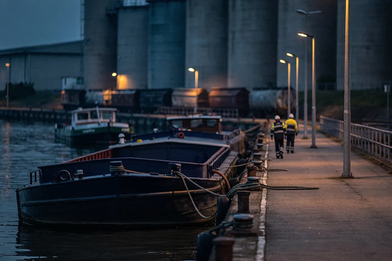 Hafenbecken zur blauen Stunde mit vertäuten Binnenschiffen, Kailampen spiegeln sich im Wasser, Silos und ein Gleisanschluss dahinter