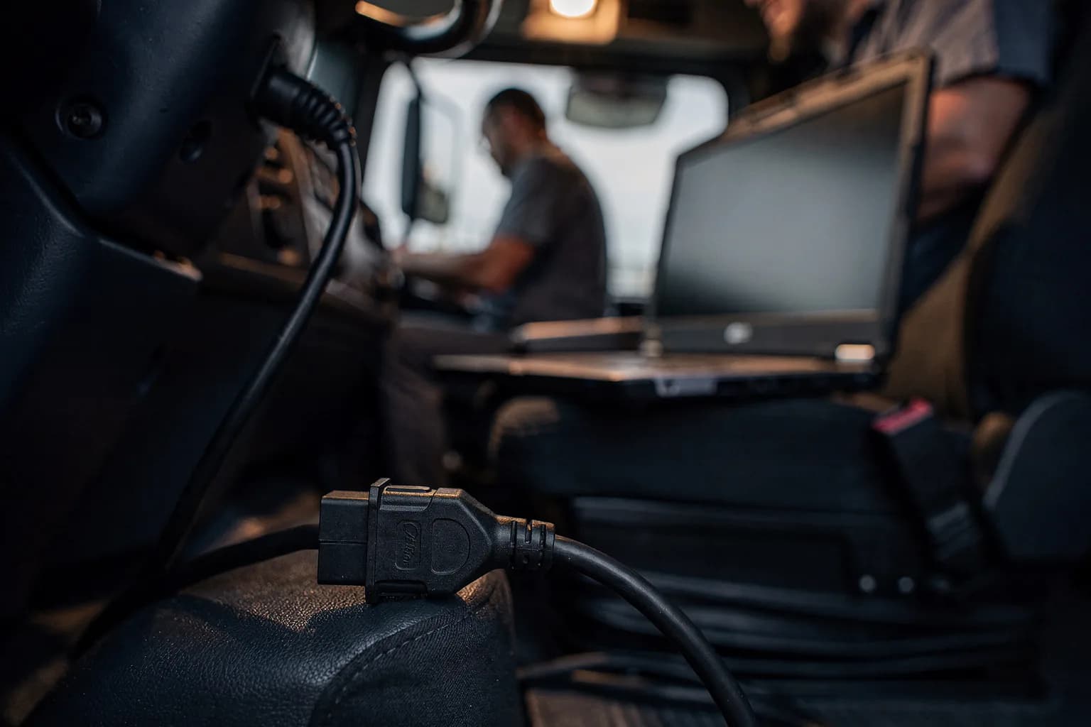 Diagnosestecker unter dem Armaturenbrett eines Lastwagens, Kabel führt zu einem robusten Laptop auf dem Sitz, warmes Licht in der Kabine