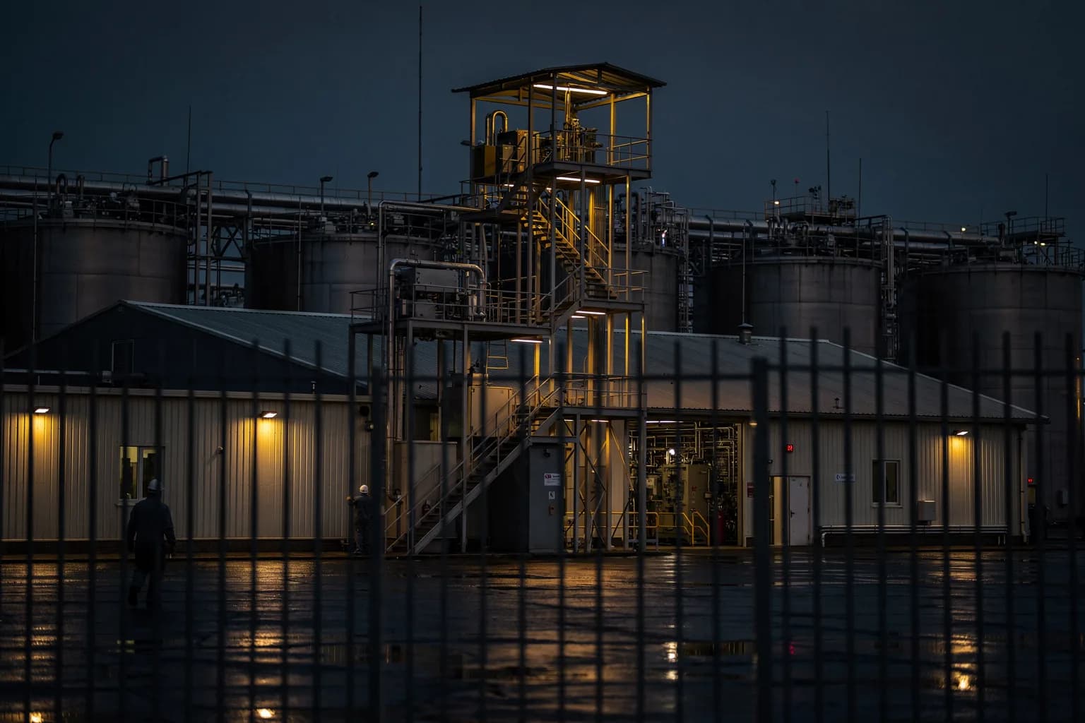 Chemiebetrieb in der Dämmerung mit Tanks und Rohrbrücken hinter einem Zaun, niedrige Gebäude, warmes Licht in einem Treppenturm