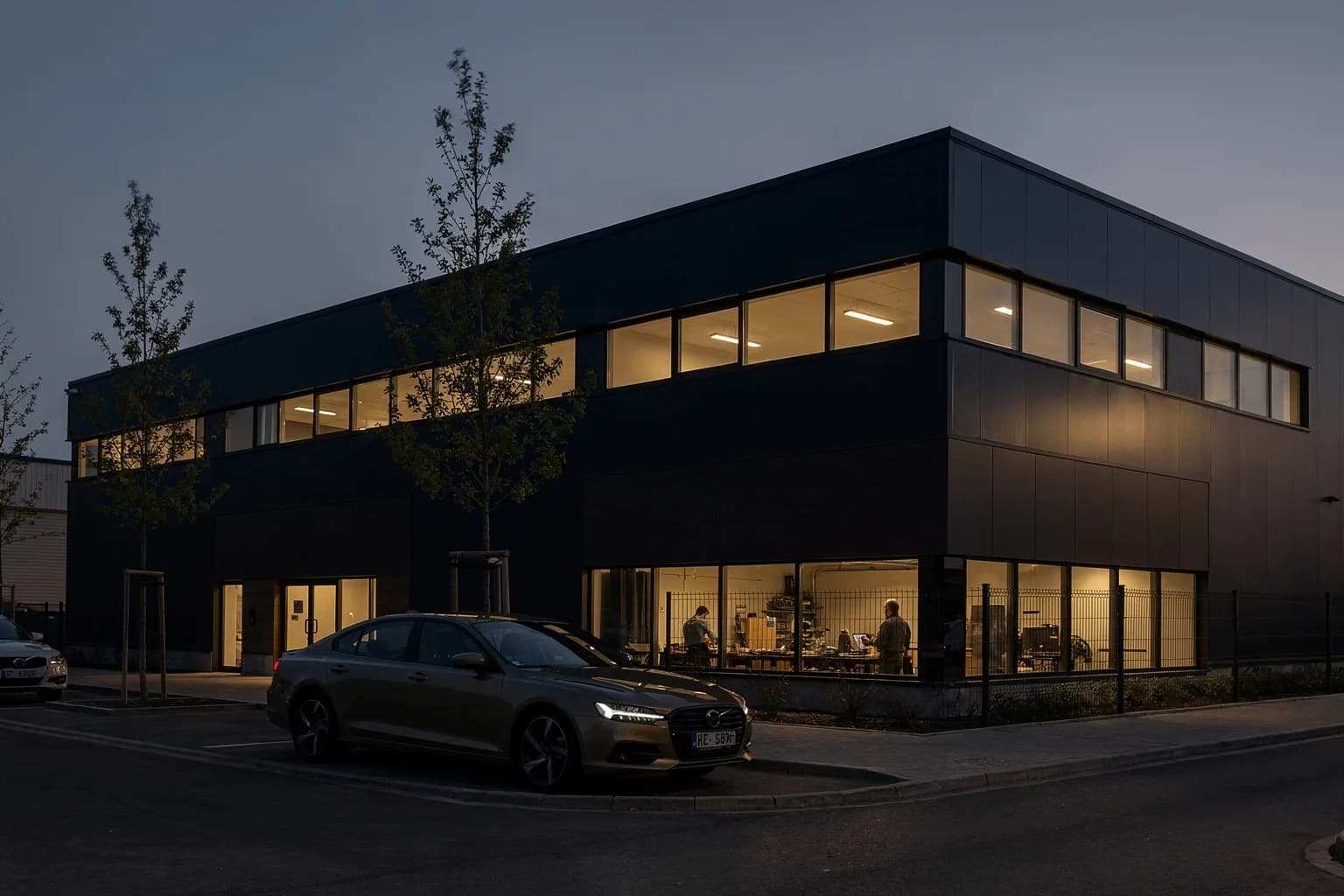 Kompaktes Industriegebäude in der Dämmerung mit beleuchteten Fensterbändern, Parkplatz und junge Bäume davor, ruhige Straße