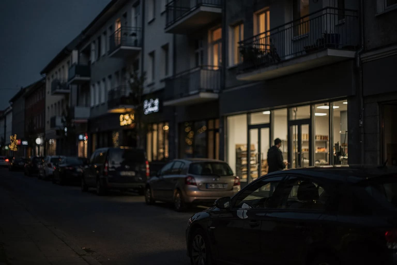 Gemischte Wohn- und Geschäftsstraße in der Dämmerung, Balkone und Ladenfronten, beleuchtete Fenster, parkende Autos