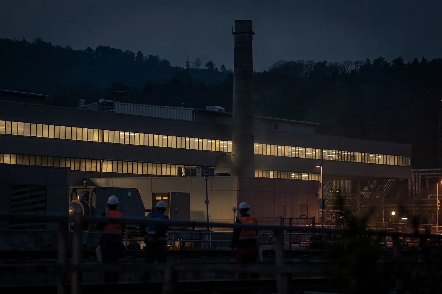 Großes Industriewerk in der Dämmerung mit langen beleuchteten Fensterbändern und hohem Schornstein, bewaldete Hänge dahinter