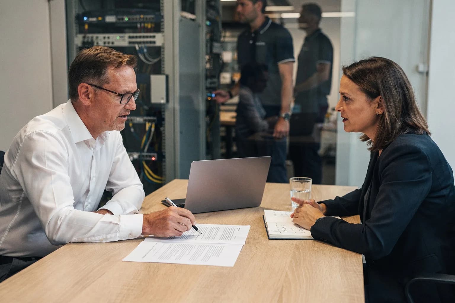 Geschäftsführer und IT-Dienstleisterin gehen an einem Besprechungstisch einen Vertrag durch, im Hintergrund ein Technikraum
