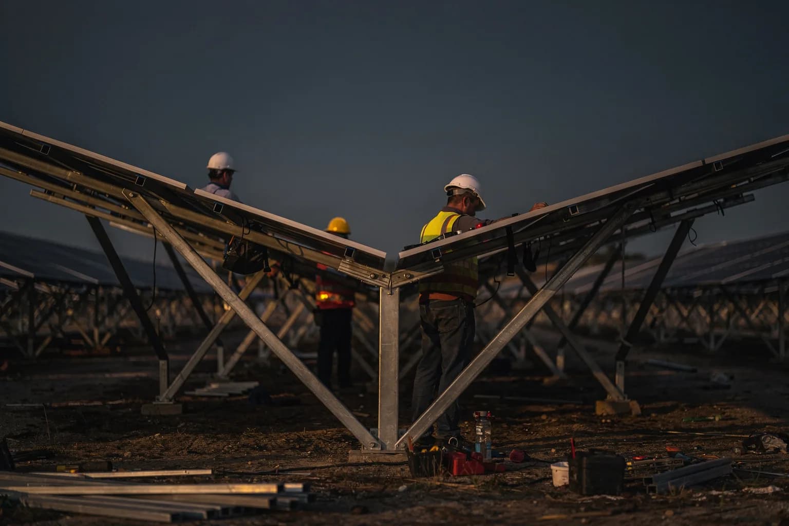 Großer Freiflächen-Solarpark im Bau, Reihen stählerner Montagegestelle, Arbeiter setzen Module ein, aus flachem Winkel in der Dämmerung aufgenommen