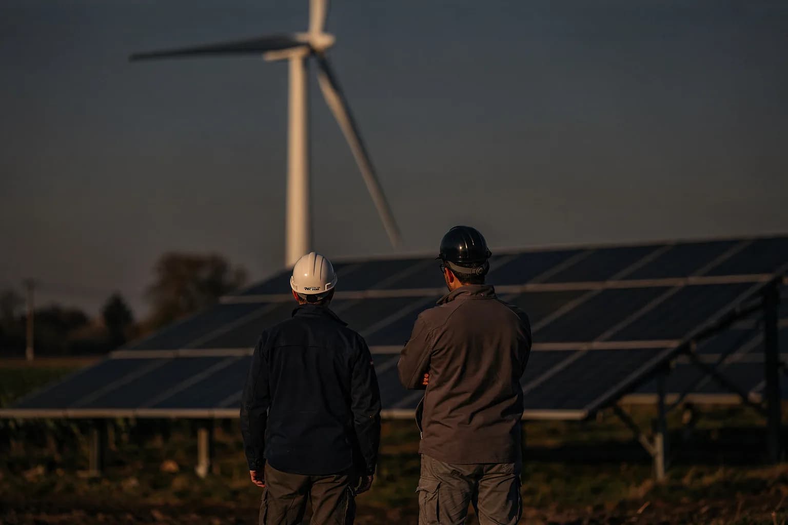 Eine Windenergieanlage und eine Freiflächen-Photovoltaikanlage am Feldrand, zwei Personen von hinten blicken hinüber, spätes Nachmittagslicht
