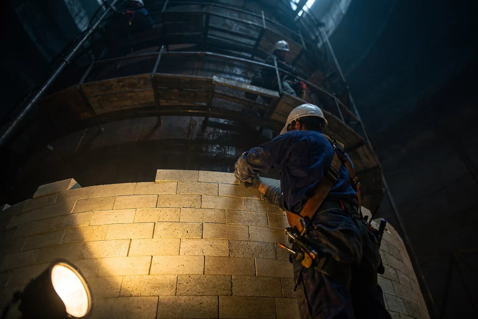 Feuerfeste Zustellung im Inneren eines großen Industriekessels, Arbeiter auf Gerüsten setzen Schamottesteine, Arbeitslicht von unten