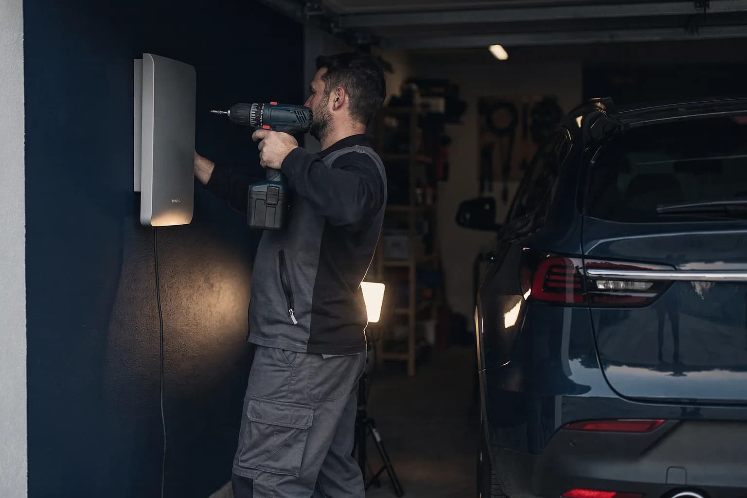 Wandladegerät wird in einer Privatgarage neben einem Auto montiert, ein Monteur von hinten mit einer Bohrmaschine, Arbeitslicht