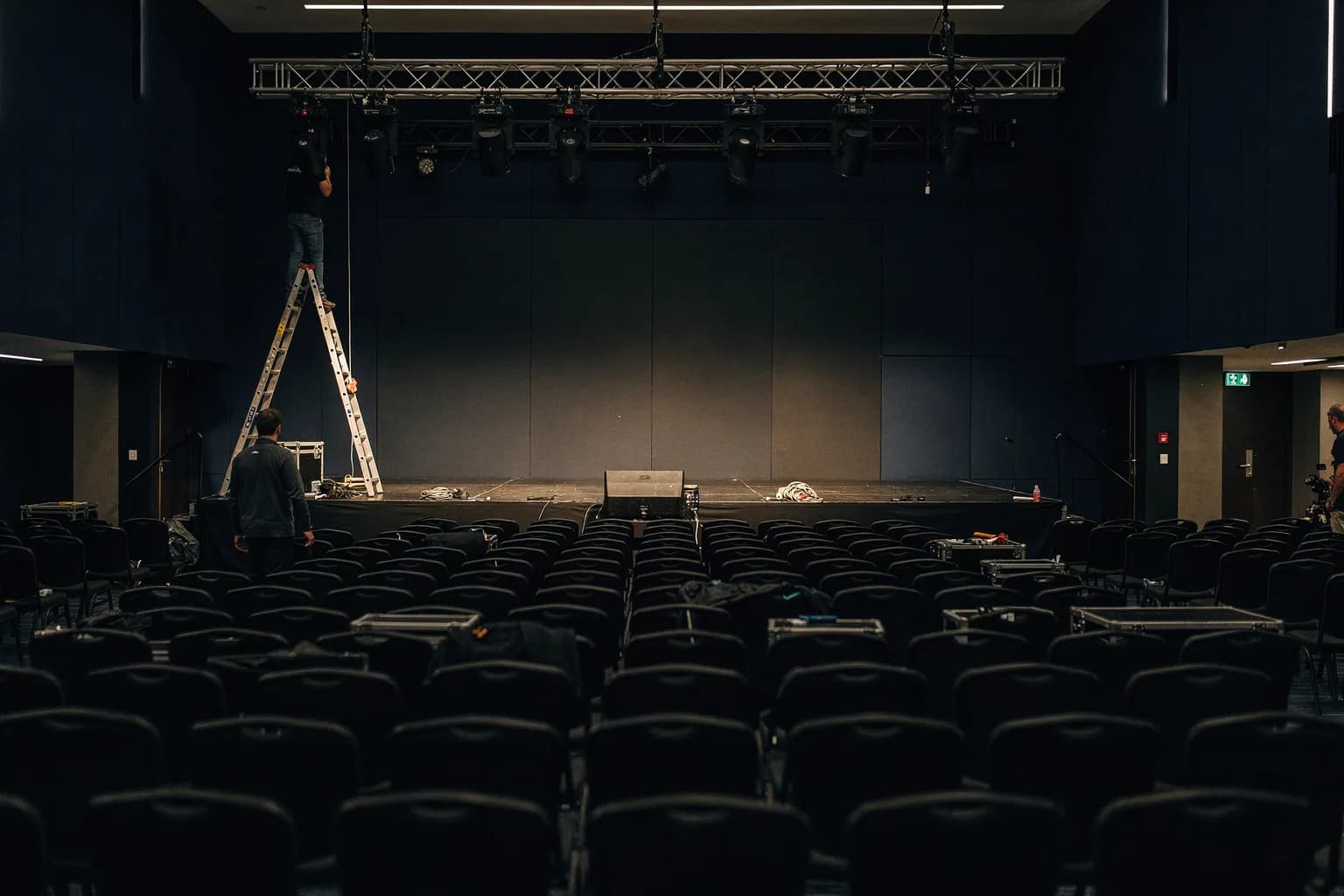 Großer Kongresssaal beim Aufbau, Reihen leerer Stühle und eine leere Bühne, ein Techniker auf einer Leiter an einem Lichttraversen-System, Arbeitslicht