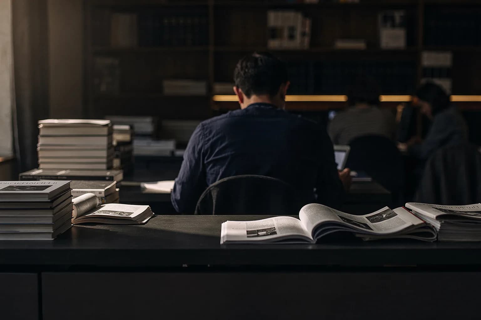 Ruhiges Verlagsbüro mit langem Schreibtisch, Stapeln von Zeitschriftenfahnen und einer Person von hinten, die eine Seite korrigiert, warmes Seitenlicht