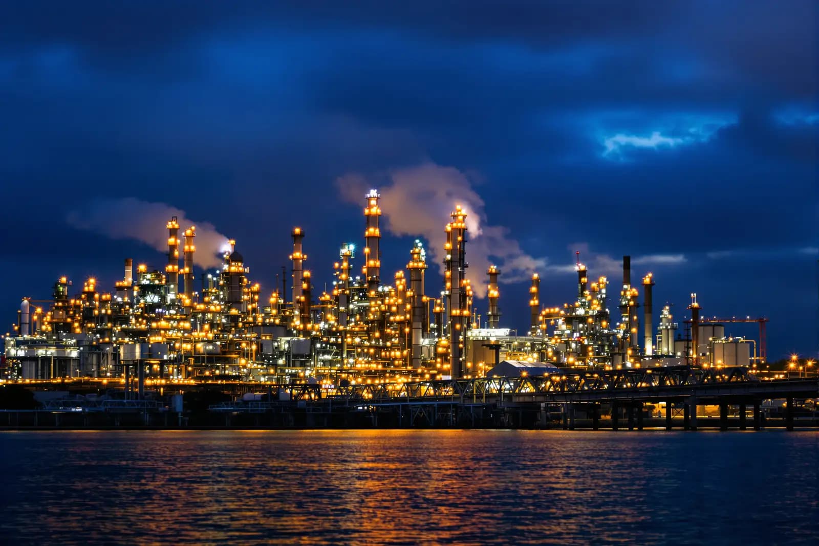 Chemieanlage jenseits eines breiten Flusses zur blauen Stunde, Kolonnen und Rohrbrücken
